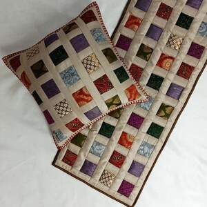 K&ouml;nnte beinhalten: Ein beigefarbener gesteppter Tischl&auml;ufer mit braunem Rand und einem passenden Kissen. Der L&auml;ufer und das Kissen sind aus Patchwork-Stoff mit verschiedenen Farben und Mustern, darunter Lila, Gr&uuml;n, Rot und Gold.