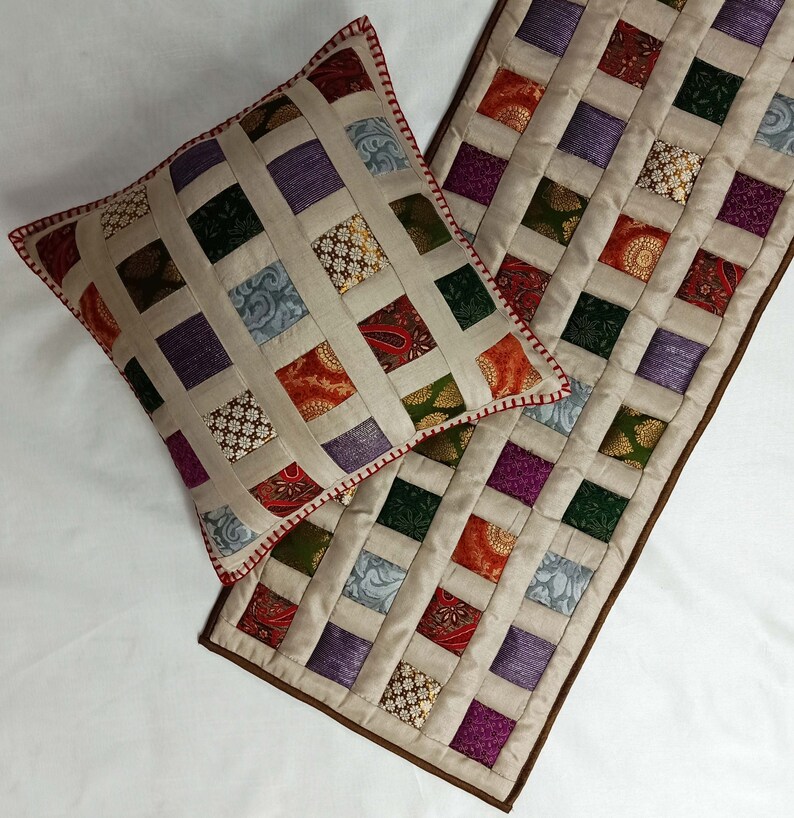 K&ouml;nnte beinhalten: Ein beigefarbener gesteppter Tischl&auml;ufer mit braunem Rand und einem passenden Kissen. Der L&auml;ufer und das Kissen sind aus Patchwork-Stoff mit verschiedenen Farben und Mustern, darunter Lila, Gr&uuml;n, Rot und Gold.