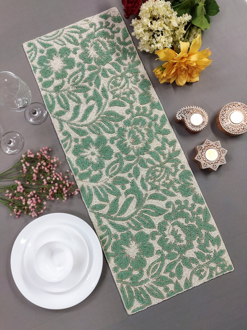 May include: A green and white beaded table runner with a floral pattern. The runner is on a gray surface with white plates and flowers.
