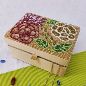 Könnte beinhalten: Eine kleine, rechteckige Box in Beige mit einem Scharnierdeckel. Der Deckel ist mit einem floralen Muster aus Perlen in Braun-, Grün-, Weiß- und Rosatönen verziert.