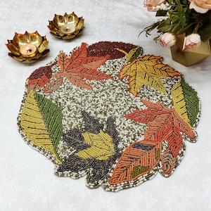 May include: A beaded placemat featuring a fall leaf design. The placemat is crafted from small beads in shades of green, brown, orange, and red. The leaves are arranged in a circular pattern.