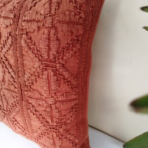 May include: Close-up of a terracotta-colored decorative pillow. The pillow features an embroidered floral pattern and vertical stitched borders. The pillow is made of a textured fabric and is a decorative home accent.