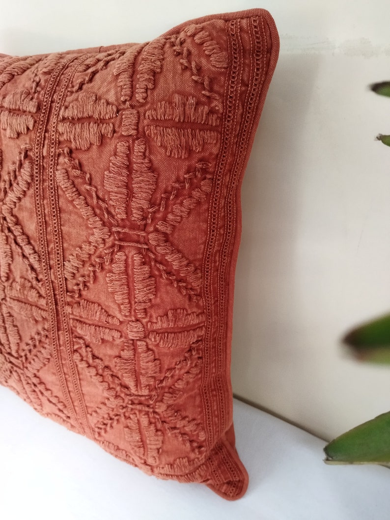 May include: Close-up of a terracotta-colored decorative pillow. The pillow features an embroidered floral pattern and vertical stitched borders. The pillow is made of a textured fabric and is a decorative home accent.