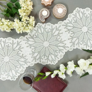 May include: A white beaded table runner with a snowflake design. The runner is made of white beads and has a silver snowflake design. The runner is on a gray surface with white flowers and candles.