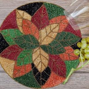 May include: Round beaded table coaster featuring a leaf design in shades of red, green, gold, and black.