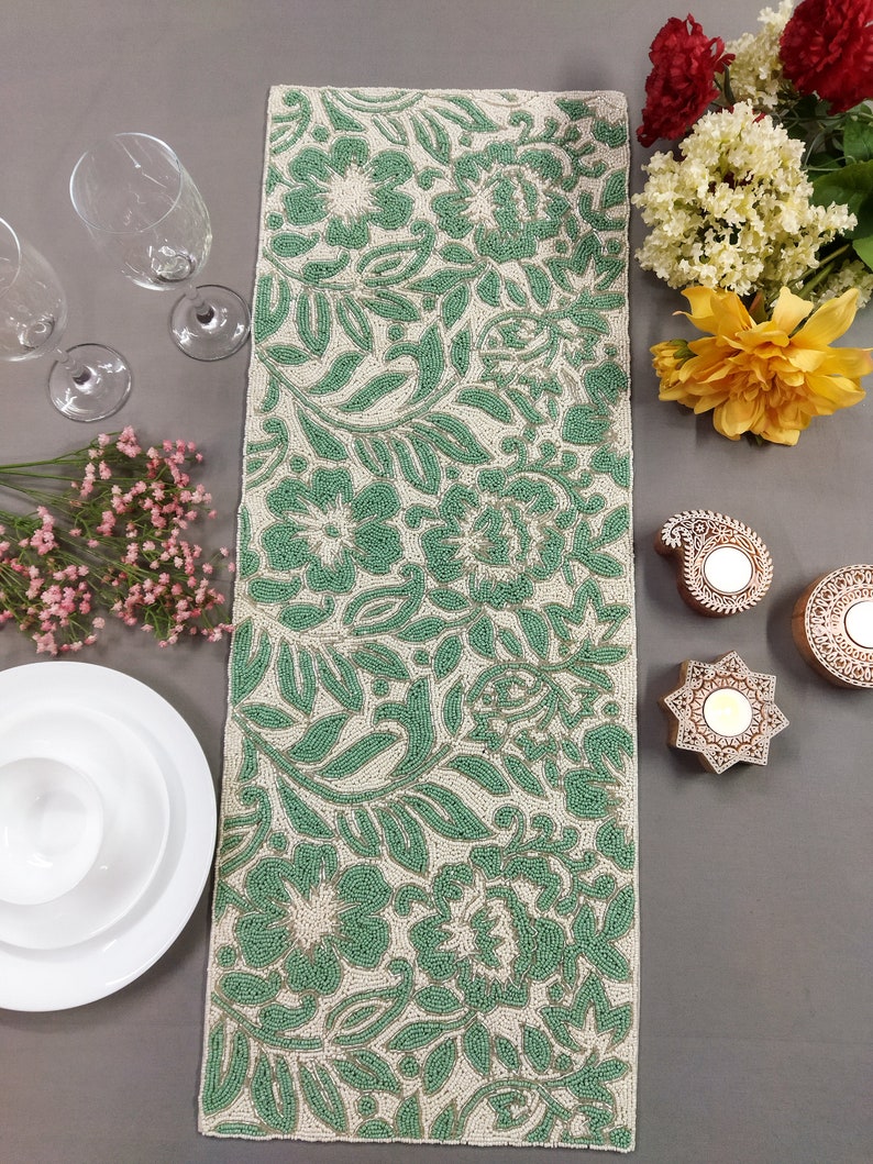 May include: A green and white beaded table runner with a floral pattern. The runner is made of small beads that are sewn together to create a raised design.