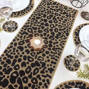 Peut inclure: Un chemin de table rectangulaire accompagné de sets de table et de sous-verres circulaires assortis, ornés d'un motif léopard en perles noires et dorées. L'ensemble comprend un bougeoir en bois en forme d'étoile avec des détails sculptés blancs.