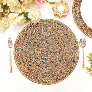 May include: A round placemat featuring a dense, concentric pattern of multicolored glass beads in shades of blue, red, green, and yellow, accented with gold-toned beads. The circular design is bordered by a thin, solid gold-toned beaded edge.