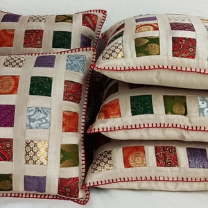 K&ouml;nnte beinhalten: Set aus vier quadratischen Kissen mit Patchwork-Muster. Die Kissen sind aus verschiedenen Stoffen in den Farben Gr&uuml;n, Lila, Orange und Gold gefertigt. Die Kissen haben einen cremefarbenen Hintergrund und einen roten, gesteppten Rand.