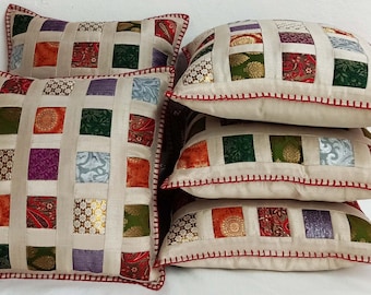Könnte beinhalten: Set aus vier quadratischen Kissen mit Patchwork-Muster. Die Kissen sind aus verschiedenen Stoffen in den Farben Grün, Lila, Orange und Gold gefertigt. Die Kissen haben einen cremefarbenen Hintergrund und einen roten, gesteppten Rand.