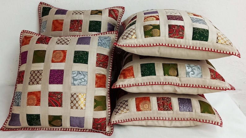 K&ouml;nnte beinhalten: Set aus vier quadratischen Kissen mit Patchwork-Muster. Die Kissen sind aus verschiedenen Stoffen in den Farben Gr&uuml;n, Lila, Orange und Gold gefertigt. Die Kissen haben einen cremefarbenen Hintergrund und einen roten, gesteppten Rand.