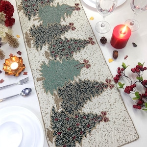 Op de afbeelding: Beaded kerstboom tafelloper. De loper heeft een crèmekleurige achtergrond met geborduurde groene kerstbomen en rode accenten. De rechthoekige loper ligt op een wit oppervlak, met borden, glazen en een brandende kaars.