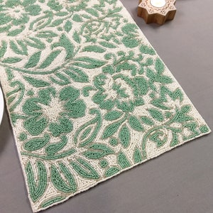 May include: A white beaded table runner with a green floral pattern. The runner is made of small, round beads that are sewn together to create a delicate and intricate design.