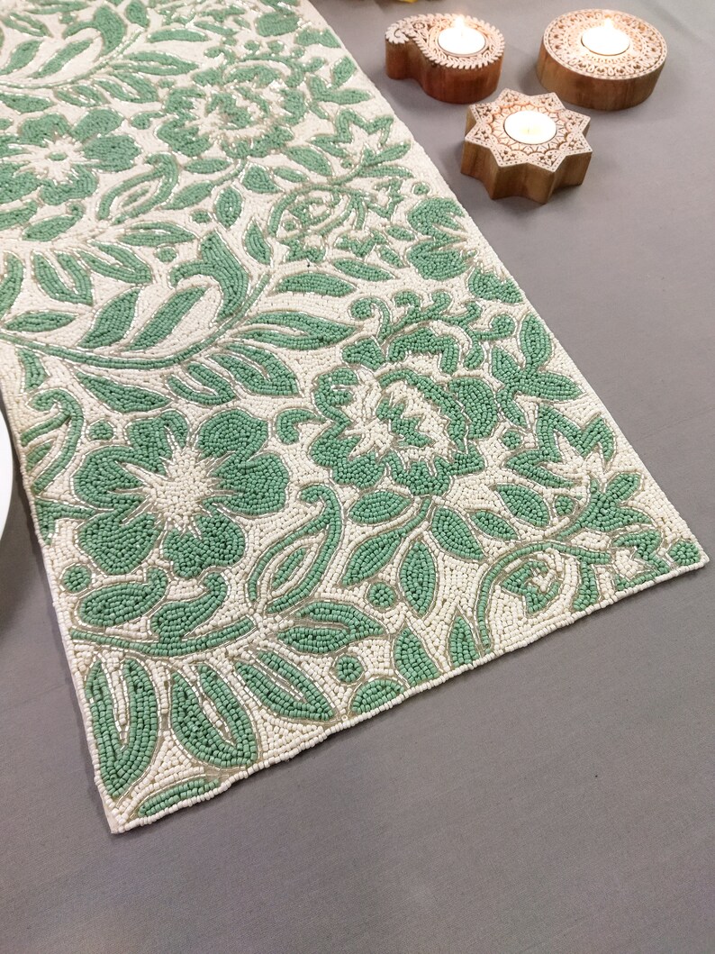 May include: A white beaded table runner with a green floral pattern. The runner is made of small, round beads that are sewn together to create a delicate and intricate design.