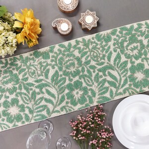 May include: A green and white beaded table runner with a floral pattern. The runner is on a gray surface with decorative items and flowers.