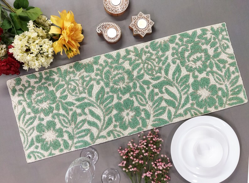 May include: A green and white beaded table runner with a floral pattern. The runner is on a gray surface with decorative items and flowers.