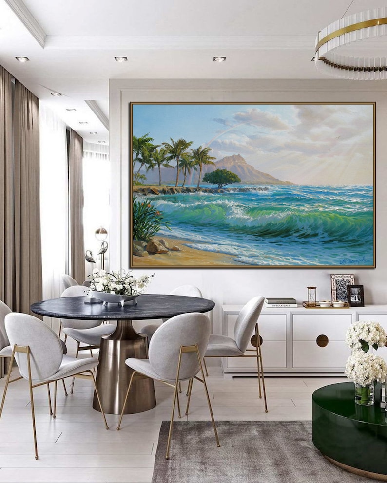 Puede incluir: Una pintura de una escena de playa tropical con palmeras, una monta&ntilde;a en la distancia y olas azules del oc&eacute;ano rompiendo en la orilla. La pintura est&aacute; enmarcada y colgada en una pared en un comedor con una mesa redonda y sillas.
