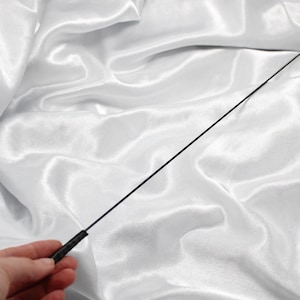 Puede incluir: Un látigo negro, delgado y flexible con un mango negro. El látigo está sobre una tela de satén blanco.