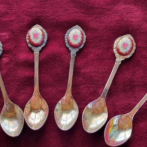 May include: Six gold-tone teaspoons with floral designs on the handles. The spoons have a white background with pink and green flowers.
