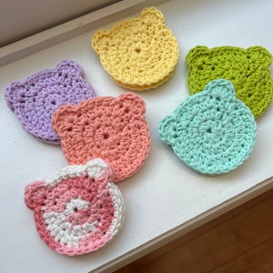 May include: Six crocheted bear-shaped coasters in various colors, including purple, pink, light blue, green, and yellow. The coasters are made with a simple stitch pattern and have a slightly raised texture.