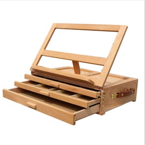 May include: A wooden easel with three drawers and a hinged top. The easel is made of light wood and has a natural finish. The drawers are open and reveal compartments for storing art supplies.