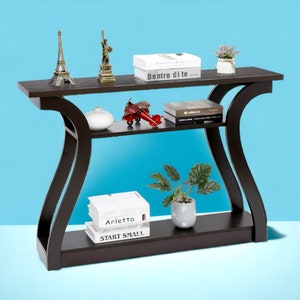 May include: A dark brown console table with two shelves. The table has a curved design and features a variety of decorative items, including a small Eiffel Tower, a statue of the Statue of Liberty, a red airplane, a white vase, a potted plant, and several books. The table is set against a light blue background.