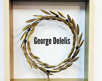 Op de afbeelding: Een gouden lauwerkrans met zwarte bessen in een lichtbeige lijst. De tekst "George Delelis" staat in zwarte letters in de lijst.