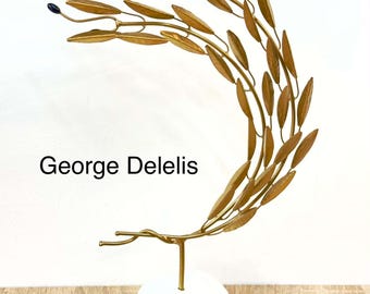 May include: A gold-colored metal sculpture shaped like a curved olive branch with small dark blue berries, mounted on a smooth white stone base. The text George Delelis is printed in black on the left side of the sculpture against a white background.