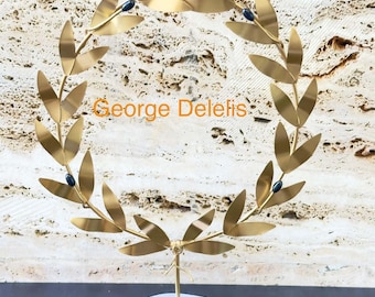 May include: A gold-colored metal laurel wreath sculpture featuring thin leaves and small dark blue accents, mounted on a smooth white stone base. The text George Delelis is printed in orange across the center of the circular wreath structure.