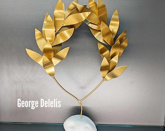 May include: A gold metal laurel wreath sculpture on a white stone base. The sculpture is a stylized representation of a traditional laurel wreath, a symbol of victory and honor. The text "George Delelis" is visible at the bottom of the image.