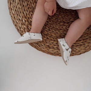 Könnte beinhalten: Babyschuhe mit floralem Muster. Die Schuhe sind hellgrau mit einem weißen und grünen Blumendesign. Die Sohlen sind hellbraun. Die Schuhe liegen auf einer gewebten, kreisförmigen Matte.