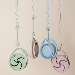 Könnte beinhalten: Vier runde Snackbehälter aus Silikon in Pastellgrün, Beige, Blau und Rosa, die jeweils an einem passenden Band mit blumenförmigen Perlen und gewellten Elementen hängen. Die Behälter haben einen flexiblen Deckel mit sternförmiger Öffnung.