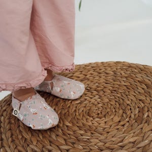 Könnte beinhalten: Babyschuhe mit hellrosa Hintergrund und einem Muster aus weißen Gänsen und grünem Laub. Die Schuhe befinden sich auf einer gewebten, kreisförmigen Matte. Die Person trägt eine hellrosa Hose.