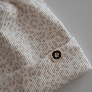Könnte beinhalten: Eine cremefarbene Rippstrick-Beanie-Mütze mit einem beigefarbenen Leopardenmuster. Die Mütze hat ein kleines Stoffetikett mit einem kreisförmigen Logo. Die Mütze ist aus einem weichen, strukturierten Material gefertigt.