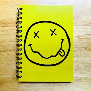 Puede incluir: Cuaderno espiral amarillo con una cara sonriente en blanco y negro con X para los ojos y la lengua fuera.