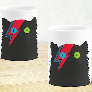 Puede incluir: Dos tazas de cerámica blanca con un diseño de gato negro. El gato tiene un diseño de rayo rojo, azul y blanco en la frente y un ojo azul y un ojo verde.