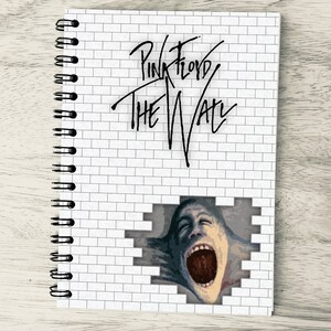 Puede incluir: Una pared de ladrillo blanco con las palabras "Pink Floyd The Wall" escritas en tinta negra. Un rostro grande y distorsionado con la boca abierta es visible a través de un agujero en la pared.