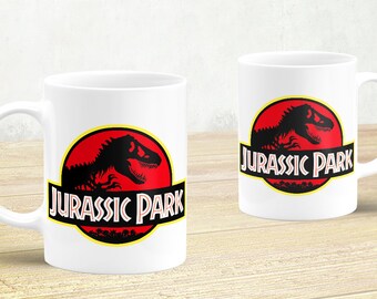 May include: Two white ceramic mugs with a black silhouette of a dinosaur and the text "Jurassic Park" in a red circle with a yellow outline.