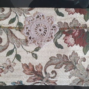 May include: A beige fabric clutch with a floral pattern and a crocheted lace embellishment.