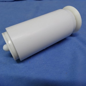 May include: A white plastic cylindrical object with a rounded base and a smaller, rounded end. The object is lying on a blue fabric surface. The cylindrical body has a smooth, glossy finish.