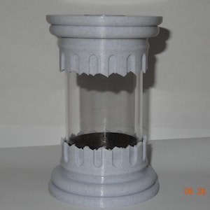 May include: A cylindrical candle holder featuring a clear glass center section held between two light gray, speckled, architectural column-style bases with arched detailing. The base has a dark circular insert with a perforated surface pattern.