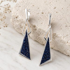 May include: Silver-coloured earrings with a geometric design. Each earring has a triangular shape filled with dark blue, textured material. The earrings are displayed on a light-coloured, textured surface.