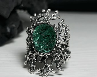 May include: A silver ring with a green stone set in a detailed, branch-like design. The ring is on a white surface.