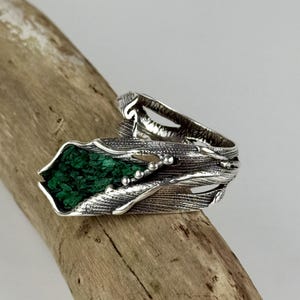 May include: A silver ring featuring a textured, branch-like metal band with a deep green, rough-cut malachite stone inlay. The metal surface displays fine, etched lines and small spherical accents, creating a detailed, organic appearance.