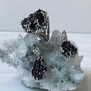 May include: A silver ring, pendant, and earrings set featuring iridescent black gemstone accents. The jewelry is displayed on a white crystal cluster.