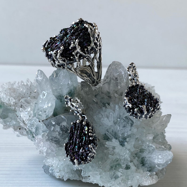 May include: A silver ring, pendant, and earrings set featuring iridescent black gemstone accents. The jewelry is displayed on a white crystal cluster.