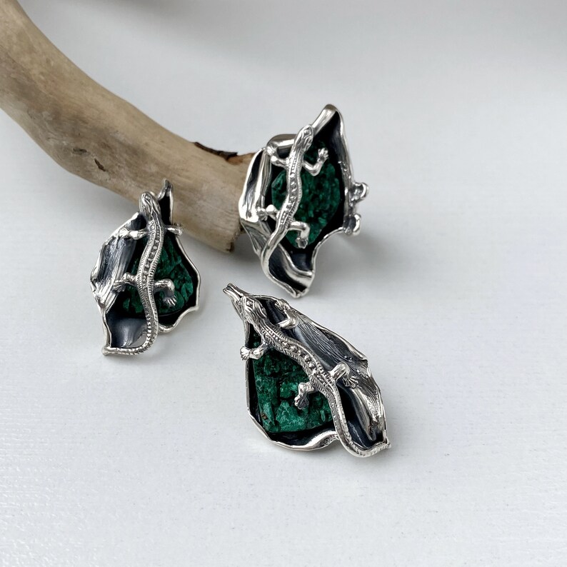 May include: A set of silver earrings and a ring featuring a green malachite stone with a silver lizard design.