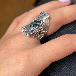 May include: A silver ring with a large, oval-shaped, iridescent gemstone set in the center. The ring has a textured, organic design with a cutout pattern.