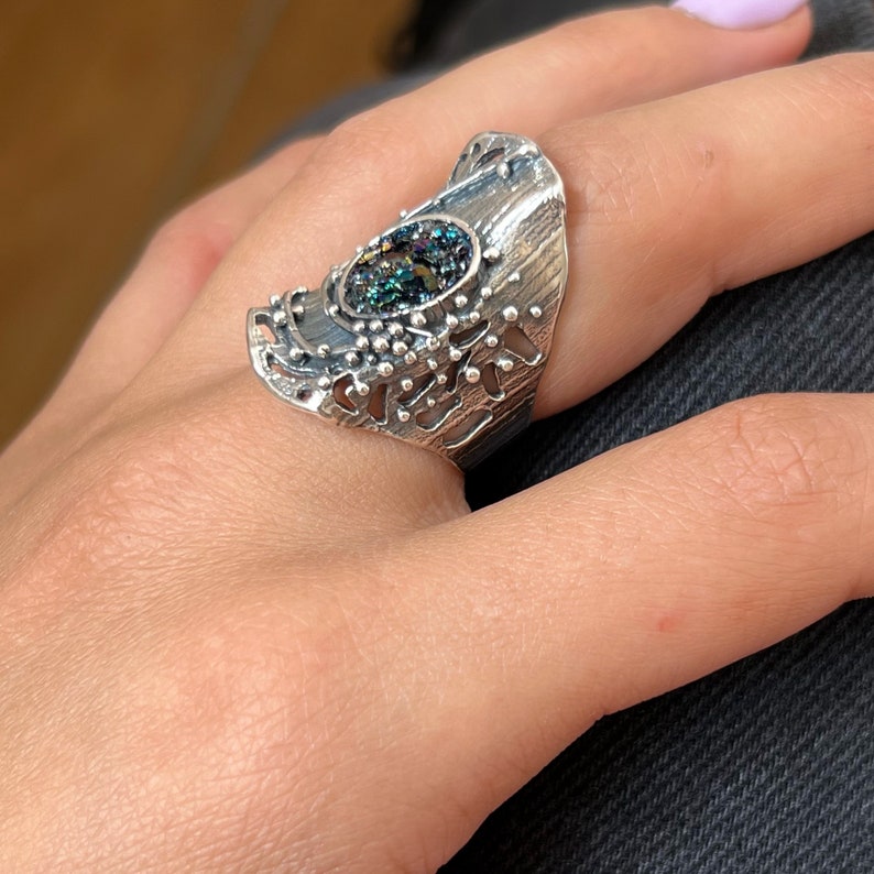 May include: A silver ring with a large, oval-shaped, iridescent gemstone set in the center. The ring has a textured, organic design with a cutout pattern.