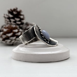 Puede incluir: Un anillo de plata con una banda texturizada y una piedra preciosa azul claro engastada en un engaste de plata. El anillo est&aacute; sobre un expositor blanco.